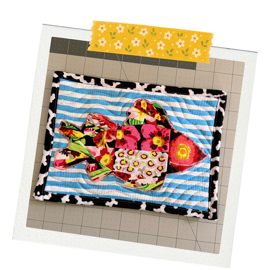 A colorful handmade quilted fabric fish ornament with floral and leopard print patterns, displayed on a gray cutting mat with a yellow floral tape strip at the top.