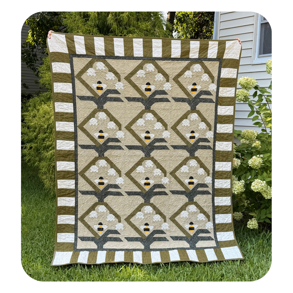 A rectangular quilted blanket or throw hanging outdoors on grass, featuring a repeating bee-and-flower pattern with olive green, cream, white, and small yellow accents. The quilt is photographed in a real yard with shrubs and house siding visible in the background.