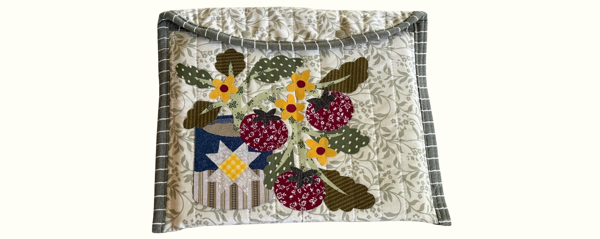 A square quilted fabric
laptop sleeve featuring a colorful applique design of red patterned tomatoes, yellow flowers, green leaves, and a blue-and-white star on a cream floral background.