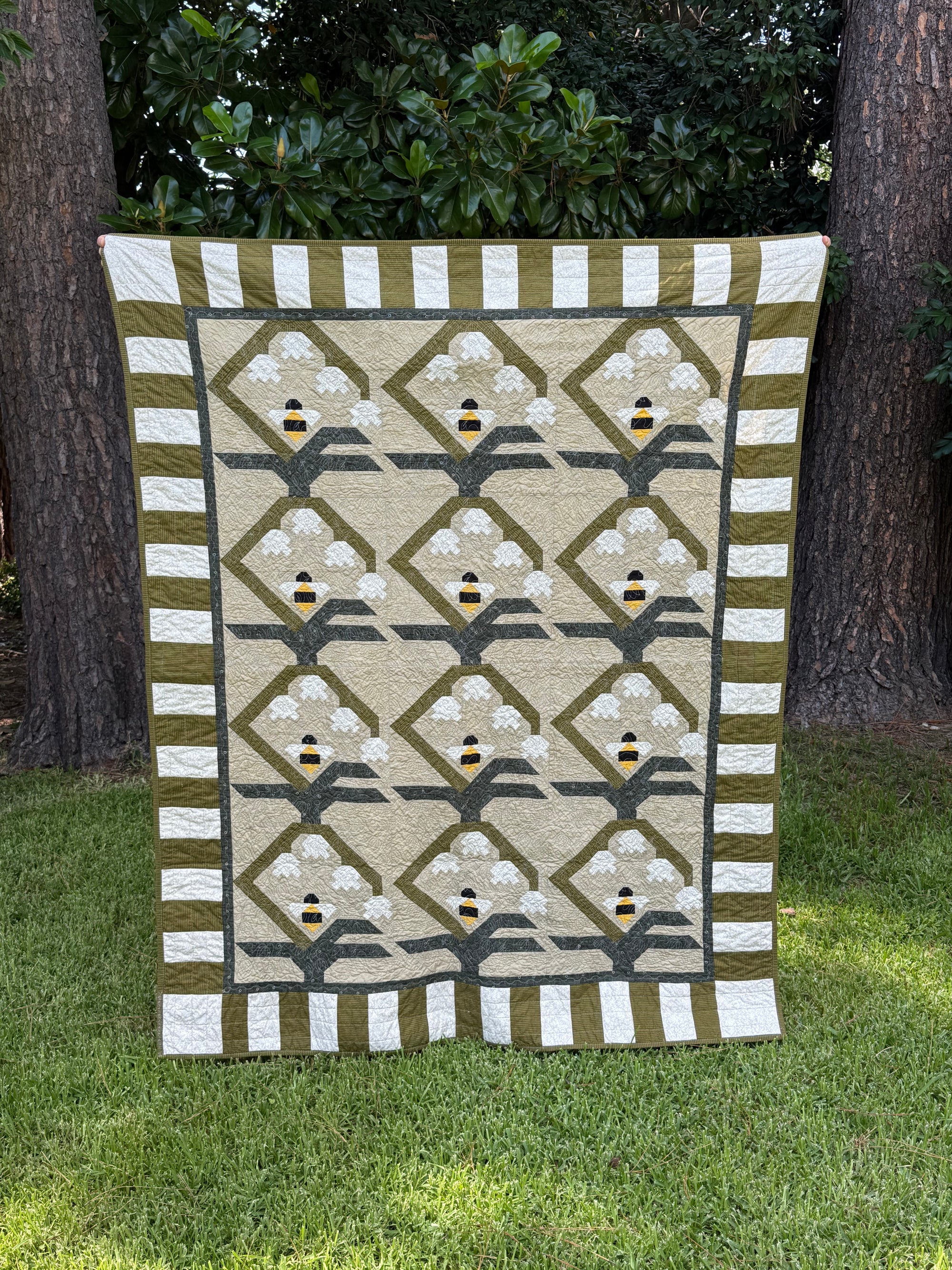 A rectangular quilted blanket or small quilt hanging outdoors on grass between two trees. The design has a beige center with repeating diamond motifs, white floral shapes, olive green borders, and small yellow-and-black bee accents.