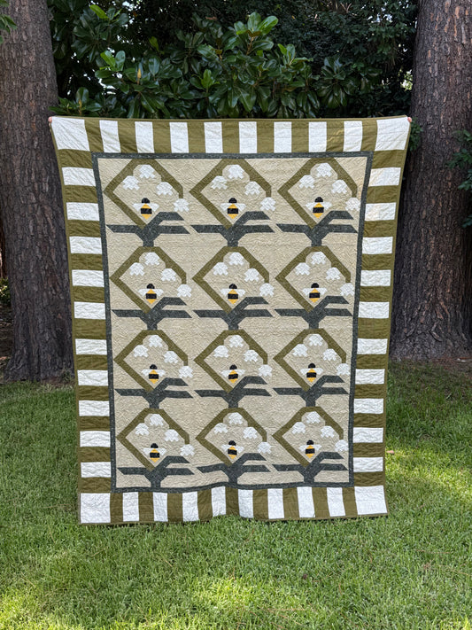 A rectangular quilted blanket or small quilt hanging outdoors on grass between two trees. The design has a beige center with repeating diamond motifs, white floral shapes, olive green borders, and small yellow-and-black bee accents.