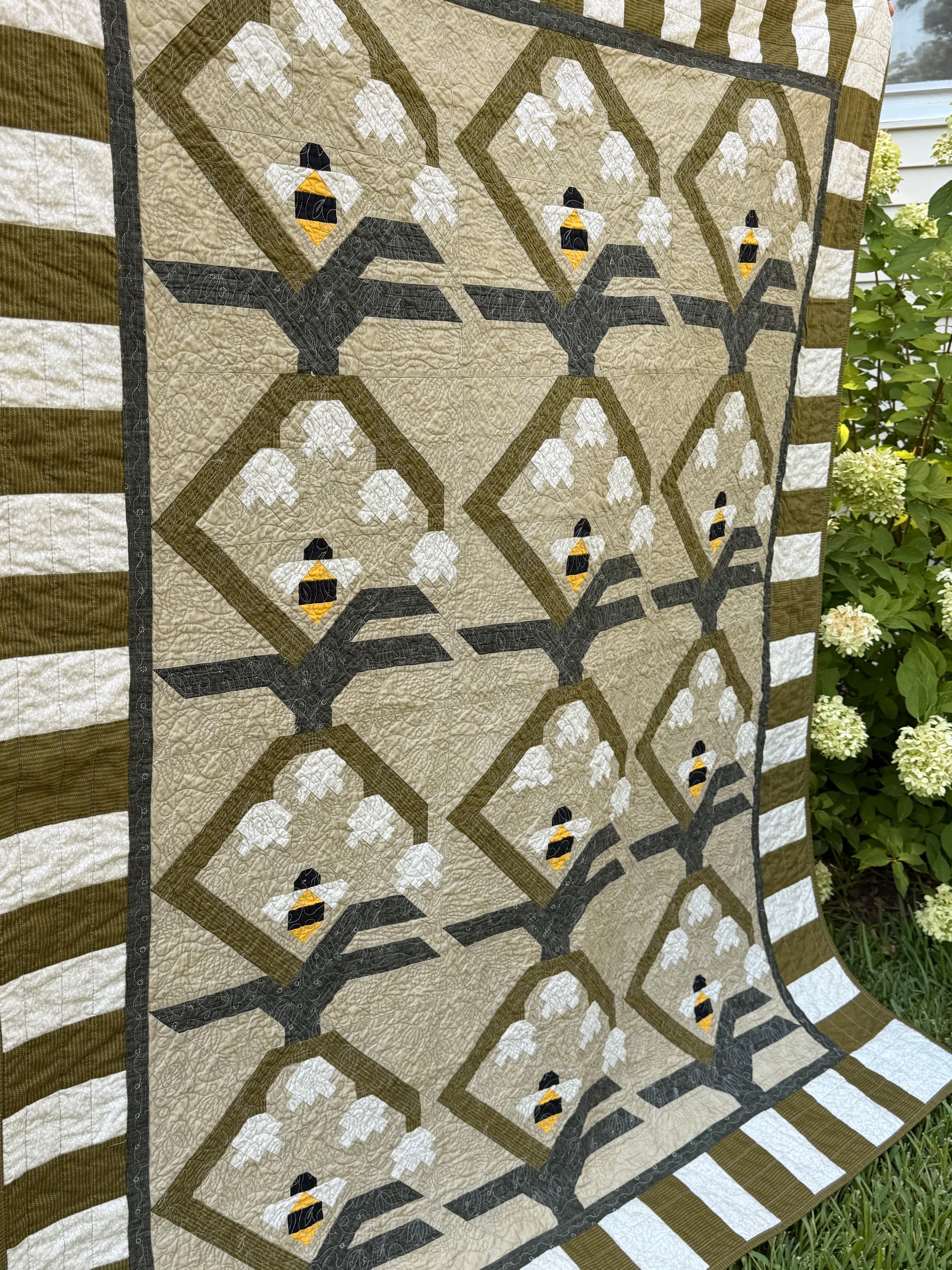 A quilted textile featuring a geometric pattern of olive green diamonds, dark gray tree branches, cream clouds, and small bumblebee motifs, displayed outdoors next to green hydrangea bushes.