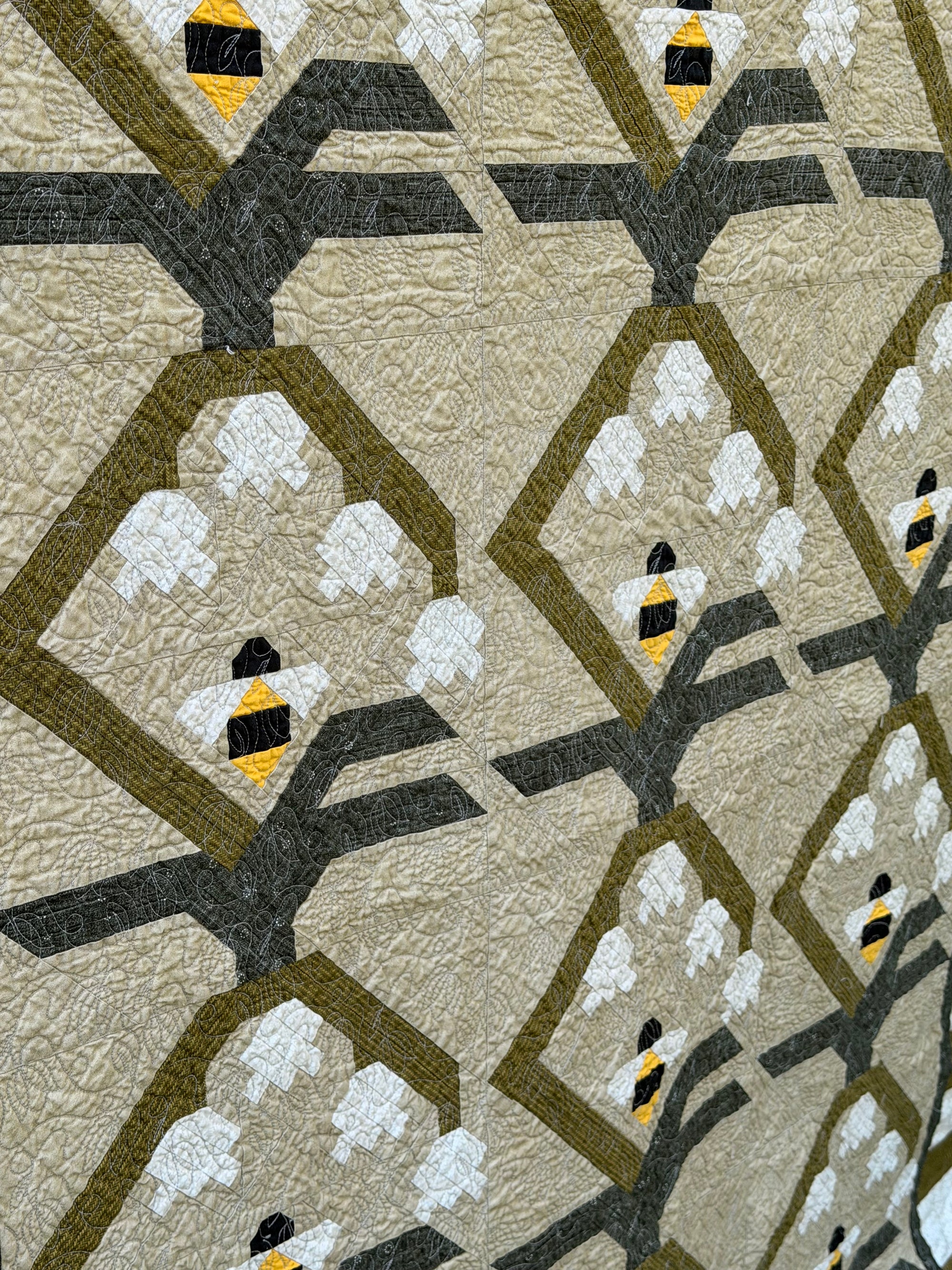 A close-up of a quilted fabric featuring a repeating pattern of stylized bumblebees in yellow, black, and white, set against olive green diamond outlines and dark gray branch-like shapes on a warm beige background.