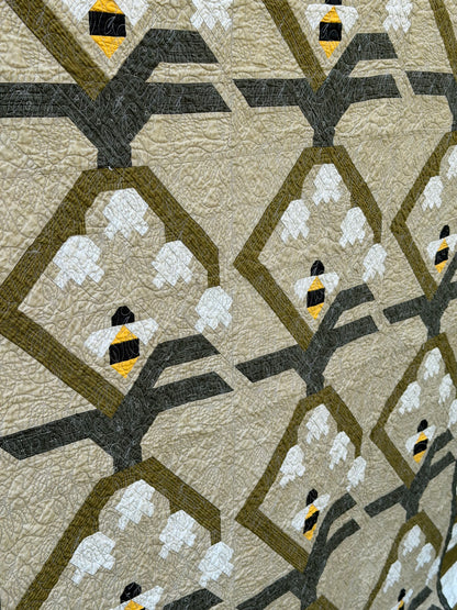 A close-up of a quilted fabric featuring a repeating pattern of stylized bumblebees in yellow, black, and white, set against olive green diamond outlines and dark gray branch-like shapes on a warm beige background.