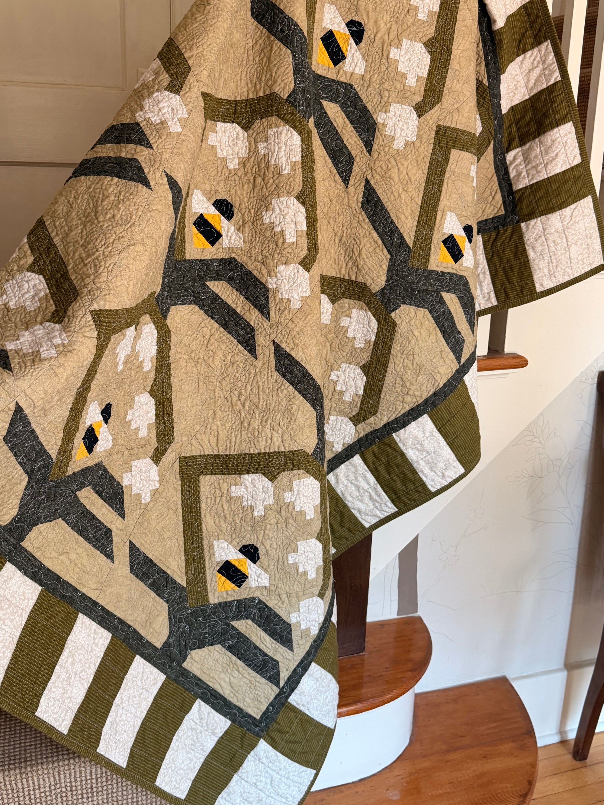 A handmade quilted textile featuring a geometric pattern of stylized bumblebees, displayed draped over a wooden staircase in a home interior.